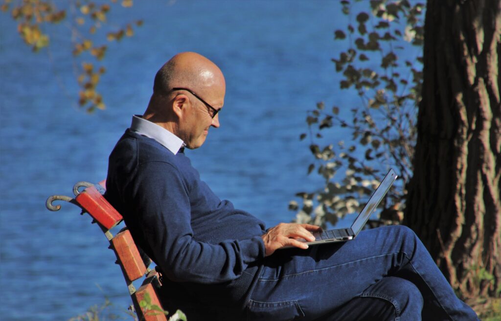 un uomo adulto seduto su una panchina in riva a un lago con un computer aperto appoggiato sulle sue gambe